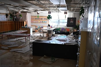 A classroom sits dark with ceiling tiles and other debris scattered all over the floor and desks
