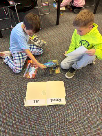 Two elementary-aged boys look at photos beside a folder that reads "Real" and "AI"