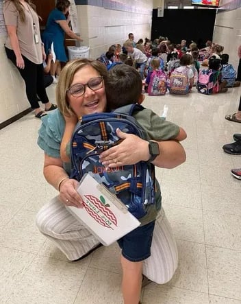 A smiling teacher with blonde hair and glasses kneels down to hug a young, male student