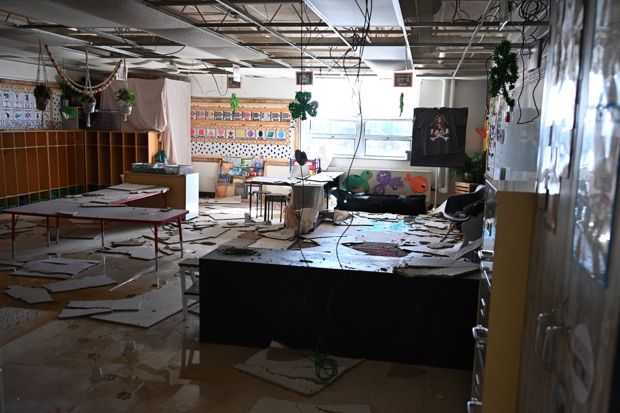 A classroom sits dark with ceiling tiles and other debris scattered all over the floor and desks