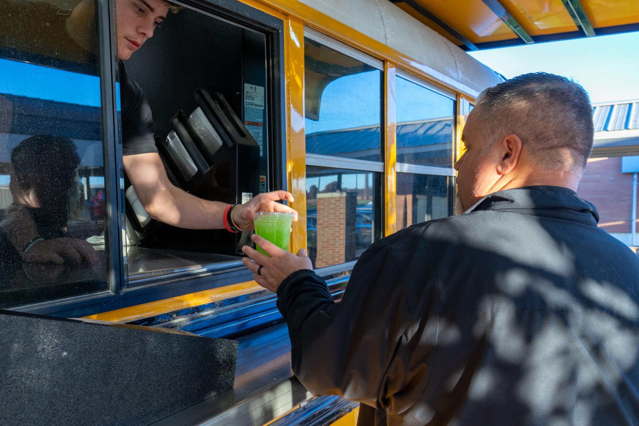 A teenage boy hands a man a drink from inside a bus
