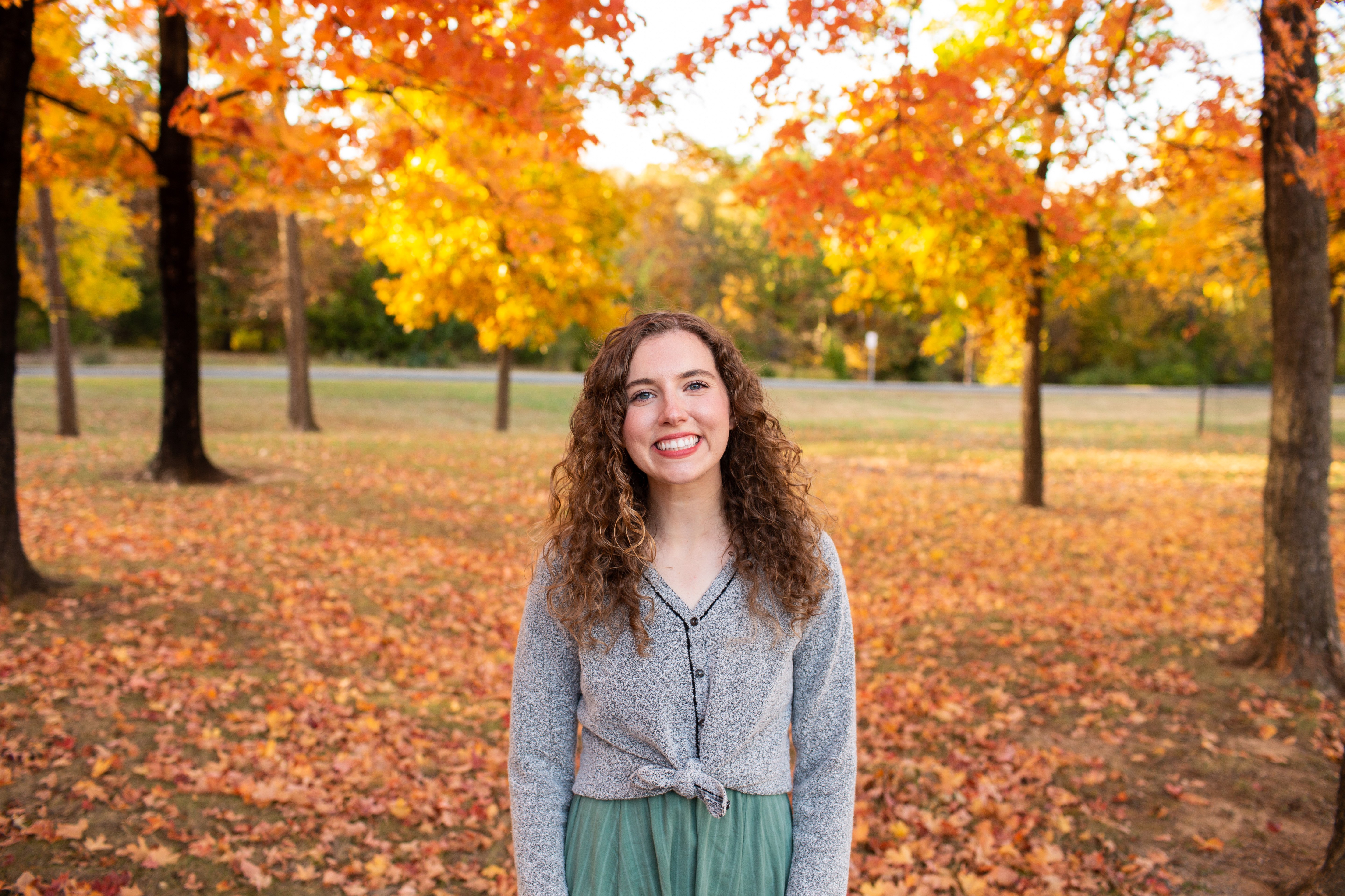 A woman in a gray sweater with brown curly hair standing outside during the fall