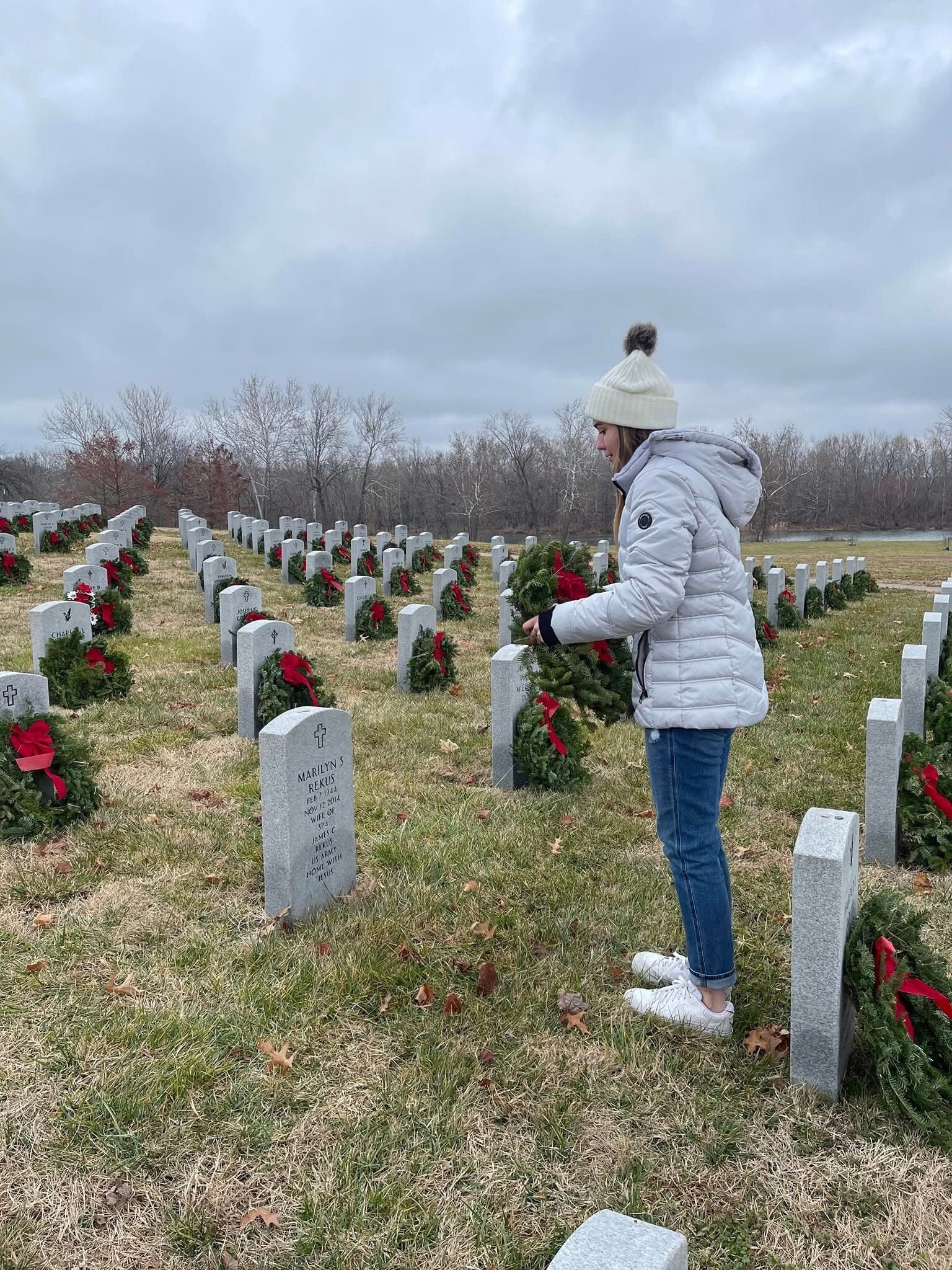 A girl holds a wreath while standing beside a headstone