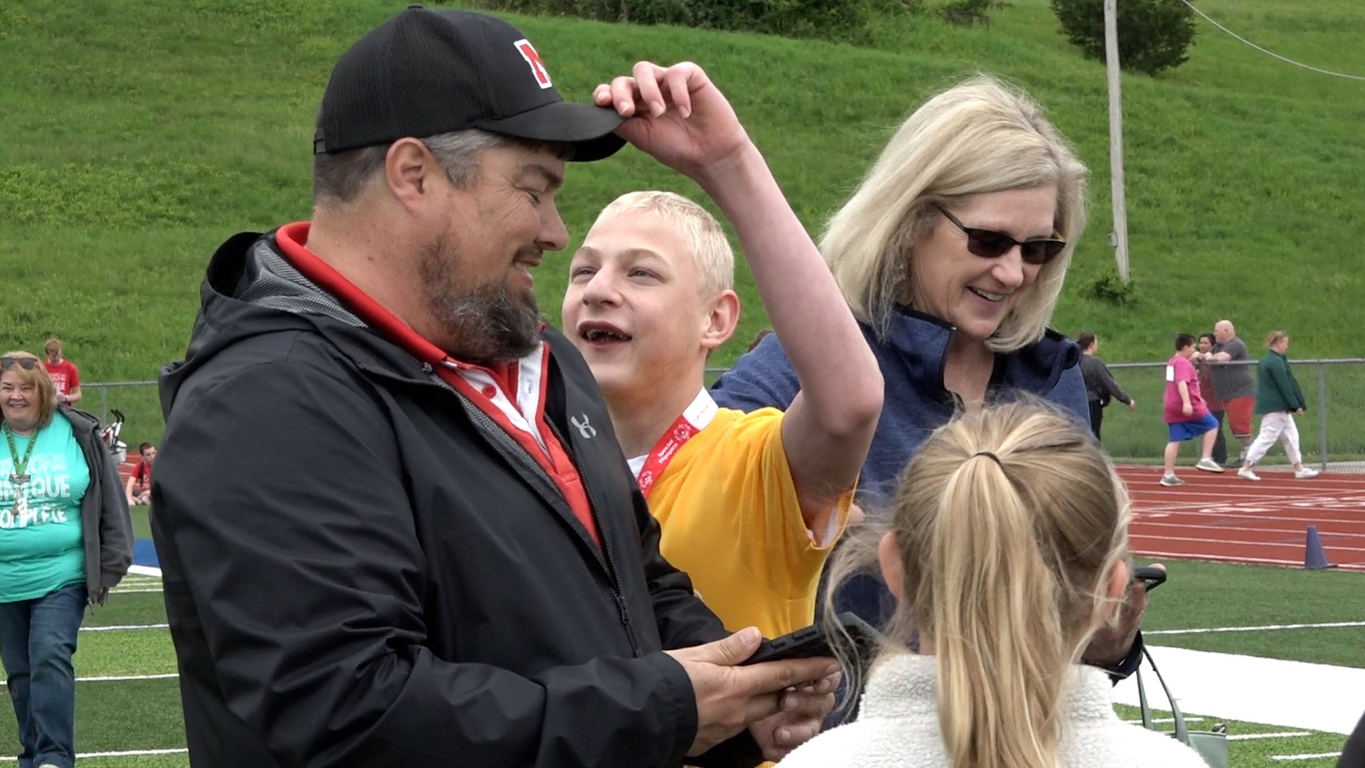 A student lifts a black hat off of a coach's head. Both have big smiles on their face