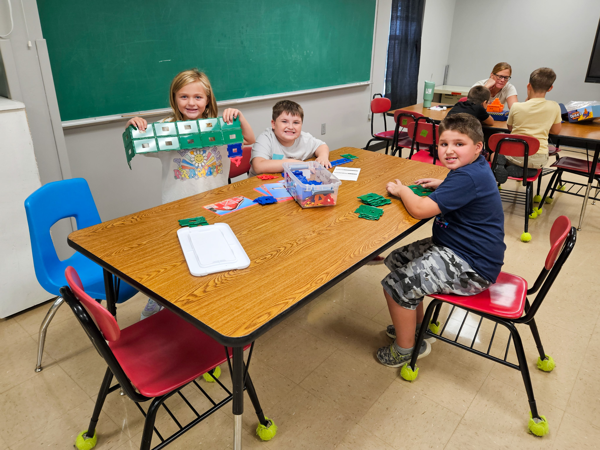 Three elementary students sit at a classroom table playing with building blocks