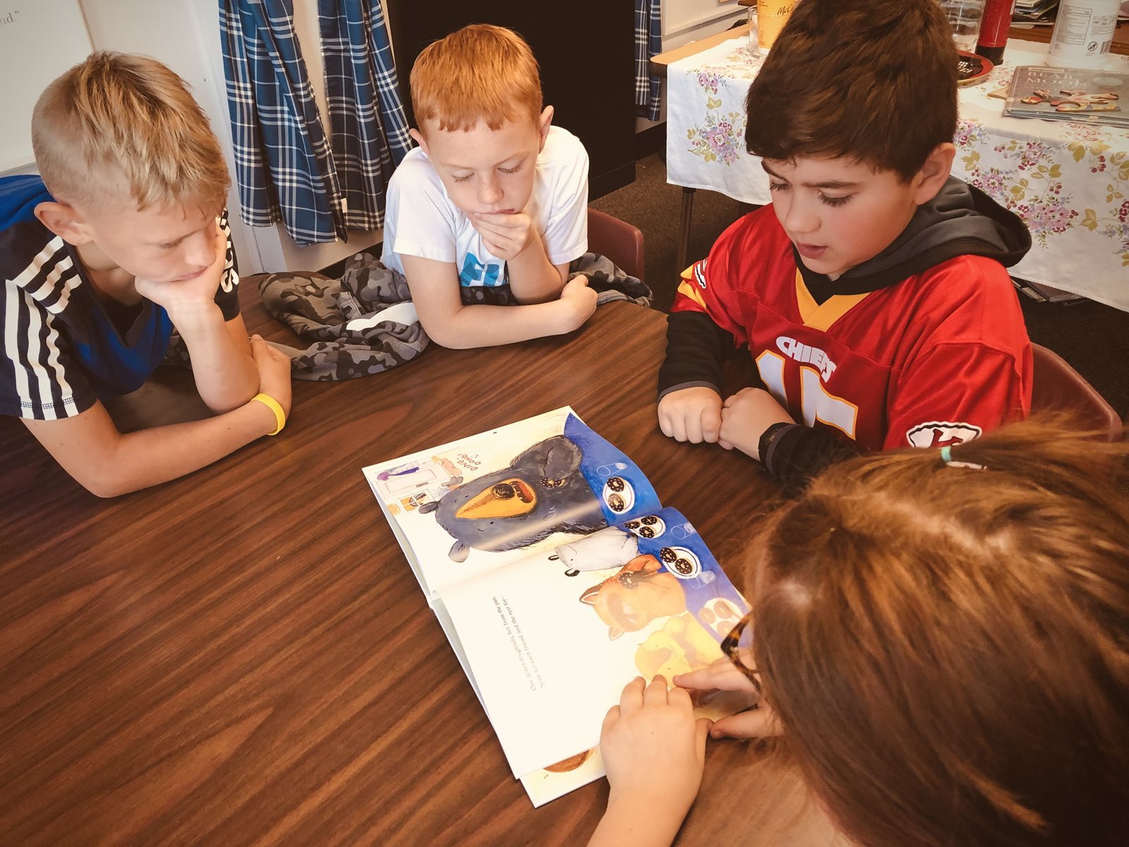 Three elementary-aged boys sit at a table and read a book