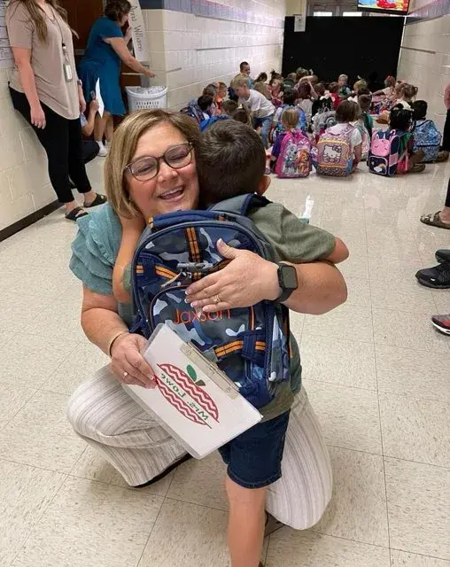 A smiling teacher with blonde hair and glasses kneels down to hug a young, male student