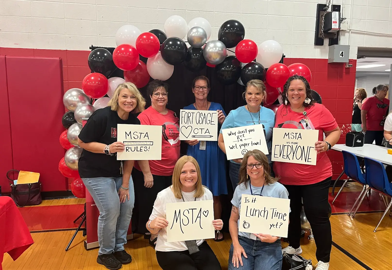 Seven female teachers pose in front of a balloon arch inside a school gymnasium with signs that read "MSTA Rules!" and "MSTA is for everyone."