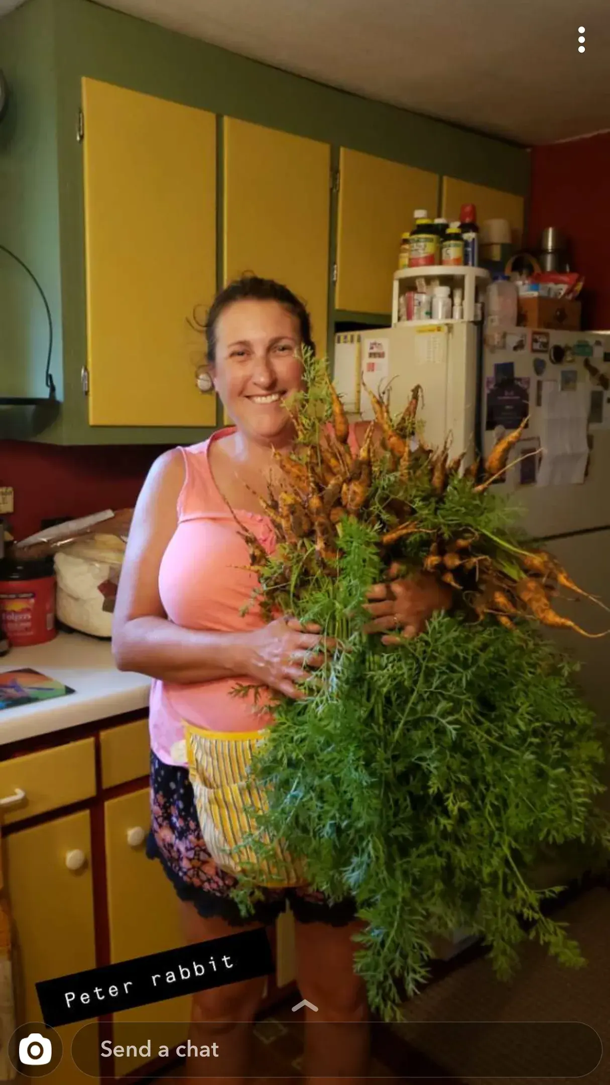 A woman in a pink tank top stands in her kitchen holding a large amount of freshly-grown carrots